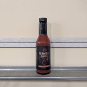 Bottle of Firewatch Sauce Co Terminal Lance hot sauce standing on a kitchen tile floor with military themed label and bold red sauce inside glass bottle